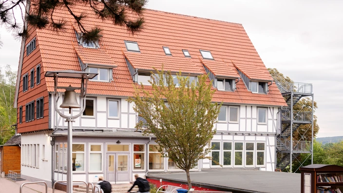 Modernes Schulgebäude mit rotem Satteldach und Fachwerk-Elementen, davor ein kleiner Baum und Fahrradständer. Das Bild vermittelt eine einladende, gepflegte Lernumgebung und aktive Schulatmosphäre.