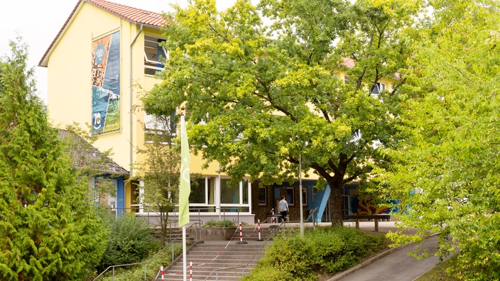 Gelbes Schulgebäude mit großem Baum im Vordergrund, umgeben von viel Grün und einem einladenden Eingangsbereich. Das Bild vermittelt eine freundliche, naturnahe Lernumgebung und einen positiven Schulstart.