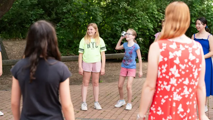Kinder und Jugendliche stehen im Kreis auf einem gepflasterten Platz im Freien und nehmen an einer Gruppenaktivität teil. Das Bild vermittelt Gemeinschaft, Spaß und soziales Lernen im Grünen.